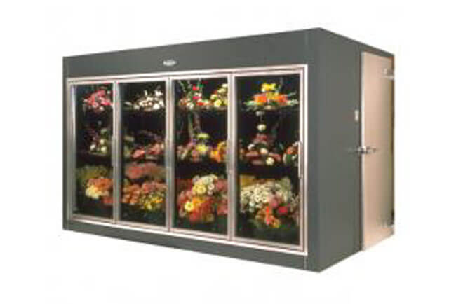 A commercial floral display cooler with a dark gray exterior, four glass sliding doors showing shelves of colorful cut flower bouquets, and a beige side access door with a handle.